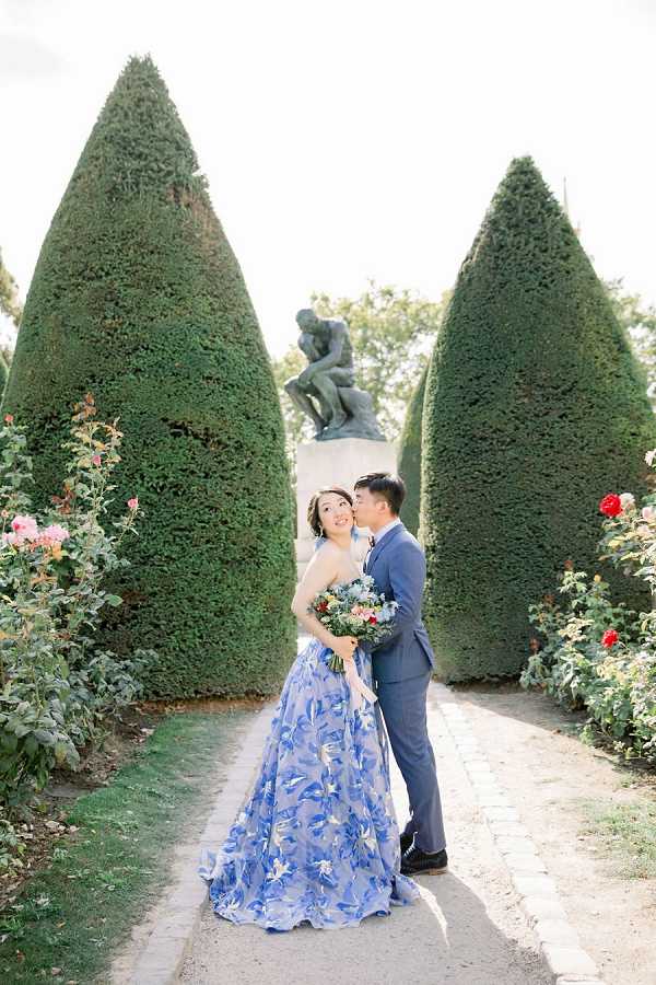 A couple poses for a portrait session outdoors in a formal French garden, identifiable by the cone-shaped topiary hedges and Rodin's 'The Thinker' sculpture visible on a stone plinth in the background. The bride wears a blue floral-patterned ball gown with a halter neckline, and the groom kisses her cheek while she smiles toward the camera; he is dressed in a slate blue suit. The bride holds a loose, garden-style bouquet featuring deep red, blush, and white blooms with greenery and eucalyptus. The composition is a full-length couple portrait taken along a gravel garden path flanked by rose bushes with pink and red blooms.