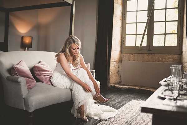A bride is getting ready indoors, seated on a light grey sofa and fastening her strappy gold heeled sandals. She is wearing a white lace wedding dress with thin straps, and her blonde hair is styled down. The room has rustic, aged stone walls with large multi-pane windows letting in warm natural light, and a dark wood floor partially covered by a patterned rug. Dusty rose velvet cushions accent the sofa, a table lamp with a warm glow is visible in the background, and a dark wood coffee table with glassware sits to the right of the frame. The overall styling combines rustic architecture with soft, muted tones. This is a medium portrait shot with a shallow depth of field.