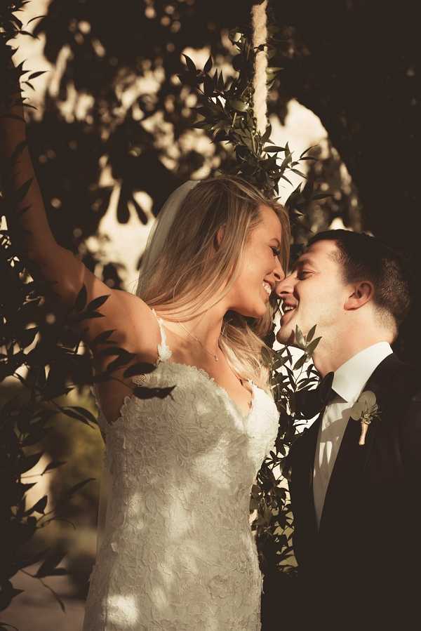 A close-up couples portrait taken outdoors, showing the bride and groom facing each other with foreheads nearly touching, both laughing. The bride wears a white lace appliqué gown with thin spaghetti straps and a veil, with long blonde hair worn down. The groom wears a dark navy or black suit with a white shirt and a small greenery boutonnière. The image is bathed in warm golden-hour light filtered through leafy branches in the foreground, creating dappled shadows across the couple. The composition is a tight portrait shot with foliage framing the subjects on all sides.