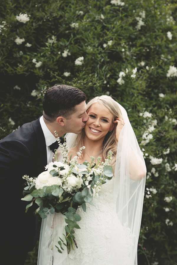 A couple portrait taken outdoors with a flowering shrub as the backdrop. The groom, wearing a navy suit with a black bow tie, kisses the bride on the cheek while she smiles broadly at the camera. The bride wears a fitted lace white gown with a long cathedral-length veil and holds a loose, garden-style bouquet of white garden roses, white wax flower, and eucalyptus with trailing greenery. The overall decor palette is white and green, consistent with a classic modern styling theme. Close-up portrait composition with shallow depth of field.