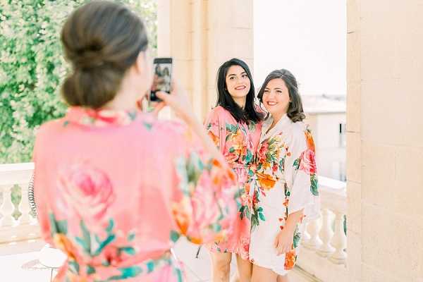 A getting-ready moment captured on an outdoor terrace or balcony with stone balustrade columns, where three women in floral satin robes are posing and taking photos on a smartphone. The woman in the foreground, seen from behind, wears a coral-pink floral robe and holds up her phone to photograph the two women facing the camera, who wear white and cream floral robes with bold orange, pink, and green botanical prints. All three have dark hair styled in loose waves or an updo. The setting appears to be a classical stone building with a balcony, likely a chateau or villa. The shot is a candid medium portrait taken in bright, soft natural daylight.