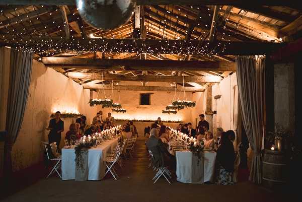 A wide-angle shot of an indoor wedding reception dinner taking place in a rustic barn or agricultural outbuilding with exposed timber beams and whitewashed stone walls. Approximately 30-40 guests are seated along two long parallel banquet tables dressed in white linen, with candlelight and greenery runners creating a warm, intimate atmosphere. The ceiling is draped with dense fairy lights and features several hanging greenery installations suspended from the beams, alongside a large mirrored disco ball near the entrance. The overall decor palette is warm amber and green, with barrel accents visible along the sides, giving the space a rustic-chic styling.