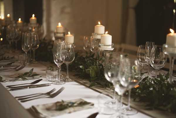 Close-up detail shot of a wedding reception table set for dinner in a dimly lit indoor venue. The table is dressed with a white linen cloth and styled with a low greenery runner composed of trailing foliage running along the center. Multiple tiered pillar candle holders with white pillar candles are spaced along the table, providing warm, low candlelight as the primary light source. Place settings include polished silver cutlery, white folded napkins with small greenery sprigs, crystal wine glasses in multiple sizes, and small tumbler glasses. The warm, candlelit atmosphere and all-green-and-white decor palette suggest a classic, understated styling approach.