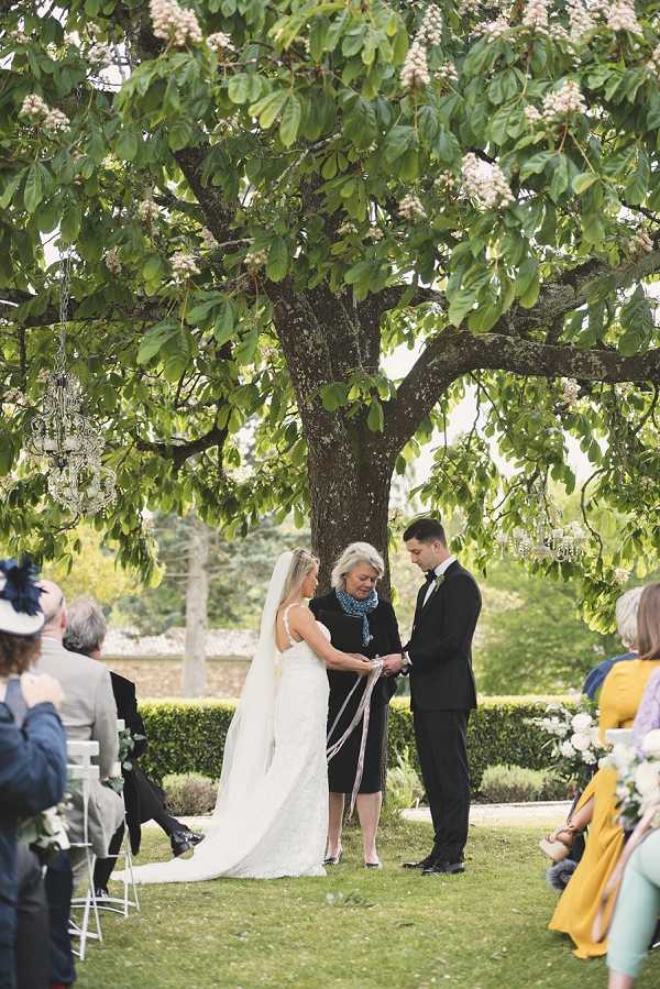 An outdoor wedding ceremony taking place beneath a large chestnut tree in a formal garden setting, with neatly trimmed hedgerows visible in the background. The bride wears a fitted ivory lace gown with spaghetti straps and a long cathedral-length veil, standing across from the groom who is dressed in a black tuxedo with a black bow tie; a female officiant in a dark dress performs a ribbon or handfasting ritual, with a white satin ribbon wrapped around the couple's joined hands. Small crystal chandeliers hang from the tree branches above the ceremony area, adding a decorative touch to the natural setting. Guests seated on white folding chairs on either side of the aisle look on, with women in mustard yellow and other colorful dresses visible; the shot is a medium-wide portrait taken from behind the seated guests, capturing the full ceremony scene.