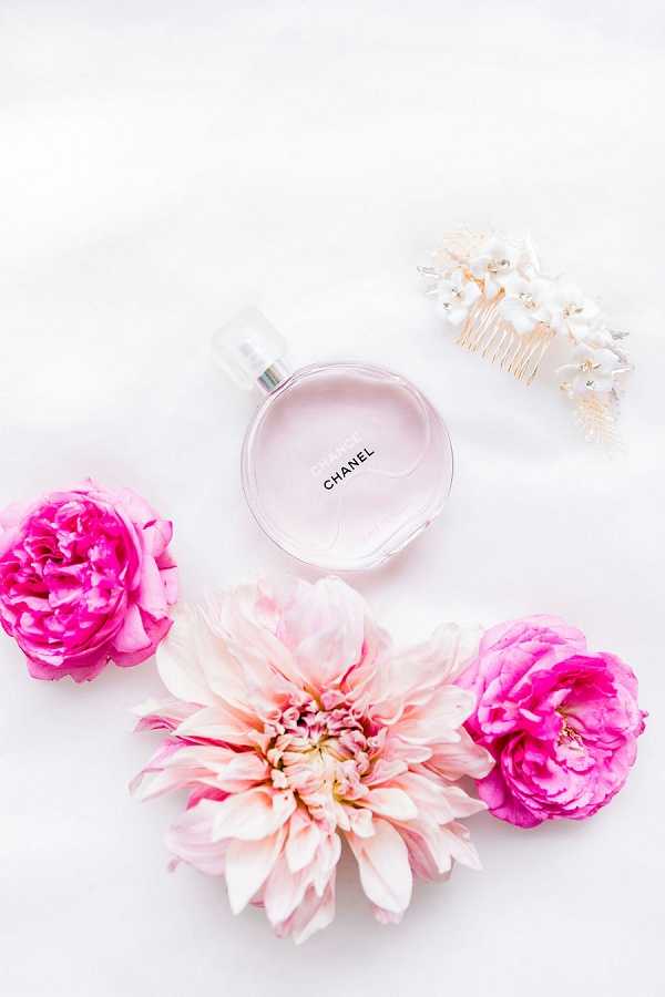 A flat lay detail shot of bridal getting-ready accessories arranged on a white fabric surface. The composition centers on a bottle of Chanel Chance Eau Tendre perfume in its signature pale pink round bottle, surrounded by three loose blooms — two hot pink garden roses and one large blush-to-cream dahlia. In the upper right corner sits a gold hair comb adorned with small white florals and pearl or crystal accents. The overall styling palette is pink and white with gold hardware, suggesting a romantic, feminine bridal aesthetic.