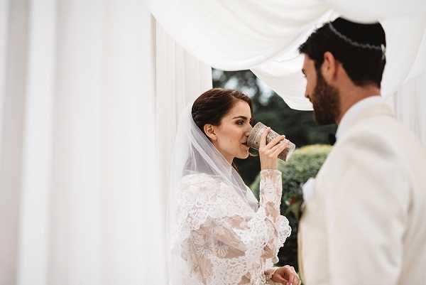 A Jewish wedding ceremony moment captured outdoors beneath a white draped chuppah, showing the bride drinking from a decorative Kiddush cup during the ritual wine blessing. The bride wears a long-sleeved lace wedding dress with a cathedral-length veil, her dark hair styled in an updo, while the groom faces her in an ivory suit and wears a white kippah. The shot is a close-up portrait framed by the white fabric draping of the chuppah, with soft natural light and a garden visible in the background. The overall styling is classic and refined, with an all-white color palette across both the bridal attire and ceremony structure.