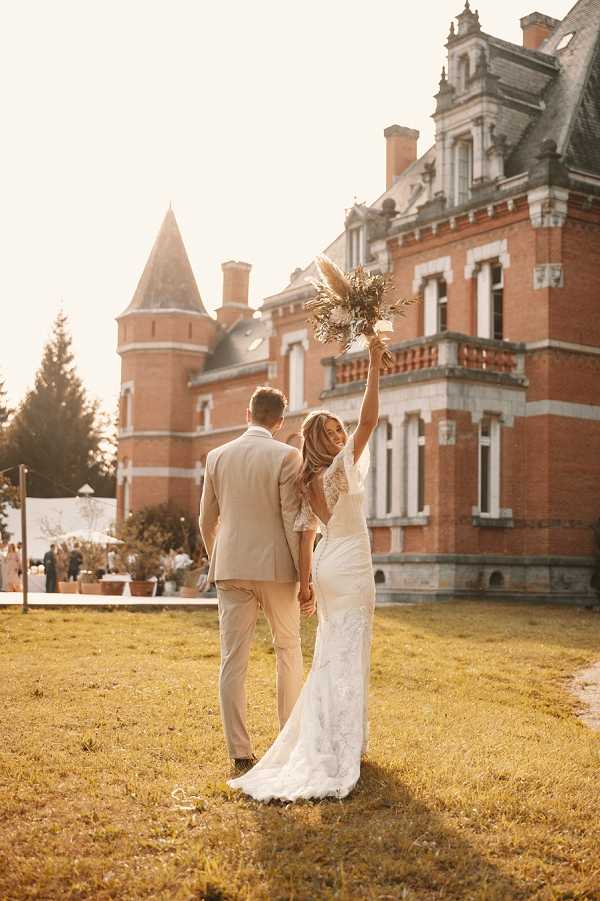 A couple portrait taken outdoors on the grounds of a red-brick French chateau with a distinctive corner tower and slate roof. The bride and groom are photographed from behind, walking together toward the chateau; the bride raises her bouquet of dried pampas grass and neutral-toned dried florals high in the air in a celebratory gesture. The bride wears a fitted lace wedding gown with a low open back and a short train, while the groom wears a light beige suit. In the background, white marquee tents and a small group of guests are visible to the left of the chateau, suggesting a cocktail hour or outdoor reception in progress. The image is shot in warm golden-hour light with a slightly hazy, sun-flared exposure, giving the overall tones an amber warmth. Full-length portrait composition. Potential venue feature image.