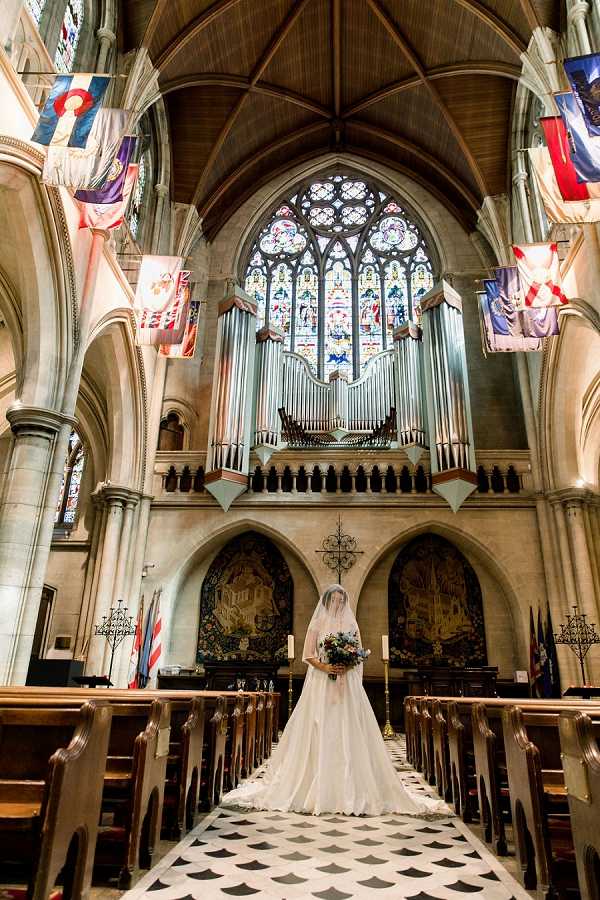 A bride stands alone at the altar end of a grand Gothic cathedral interior, facing the camera in a full-length white ball gown with a long cathedral veil and a trailing skirt. She holds a large, richly colored bridal bouquet with deep jewel tones. The cathedral features soaring ribbed vaulted ceilings, a large ornate stained glass window above a prominent pipe organ, decorative tapestries flanking the altar area, and multiple heraldic banners and flags hanging from the upper nave walls. The black and white diamond-patterned marble aisle floor leads from the wooden pew rows to the bride's position. The composition is a wide shot taken from the back of the nave, placing the bride small within the vast architectural space. Potential venue feature image.