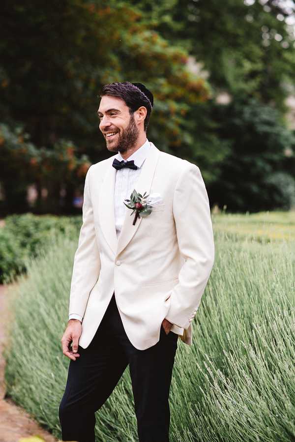 A portrait of a groom standing outdoors in a garden setting, smiling and looking to one side. He is wearing a cream dinner jacket, black trousers, a black bow tie, and a kippah, with a boutonniere featuring a white bloom, small red accents, and greenery on his lapel. Lavender plants are visible in the foreground and background, with trees softly out of focus behind him. Medium portrait shot with a shallow depth of field.
