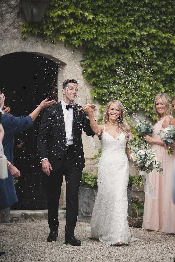 A confetti exit shot taken outdoors at what appears to be a chateau or manor, with an ivy-covered stone archway doorway as the backdrop. The couple walks hand-in-hand through a shower of white and pale pink confetti thrown by guests lining either side; the groom wears a classic black tuxedo with a black bow tie, and the bride wears a fitted ivory lace spaghetti-strap gown and carries a white and green bouquet featuring what appear to be white roses and eucalyptus. At least two bridesmaids are visible on the right in floor-length blush pink dresses, one holding a matching white and eucalyptus bouquet. The image is a medium portrait-style shot with a gravel courtyard underfoot and guests partially visible on both sides of the frame.