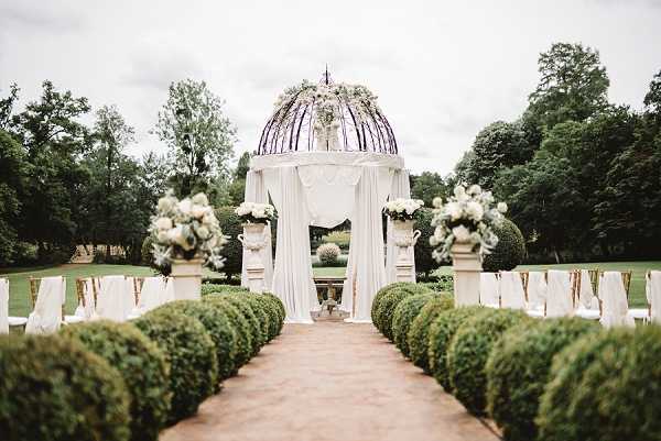 An outdoor wedding ceremony setup photographed from the end of the aisle in a wide shot, with no people present. The ceremony space is arranged in a formal garden setting featuring a central aisle flanked by trimmed boxwood spheres and white chairs with gold accents tied with ribbon. The altar is a circular domed gazebo structure with a glass or metal dome top adorned with white and green floral arrangements, draped with flowing white fabric curtains. Stone pedestals on either side of the gazebo hold large arrangements of white roses and greenery. The overall decor palette is white and ivory with gold accents, giving a classic formal aesthetic. Potential venue feature image.