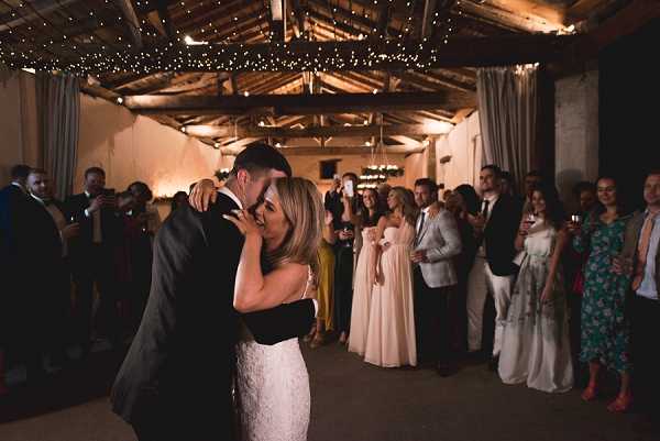 The bride and groom share their first dance in the center of a rustic indoor barn venue with exposed wooden beam ceiling draped in fairy lights and warm ambient wall lighting. The bride wears a fitted white lace gown with thin straps, and the groom is in a dark suit; the two are embracing closely with foreheads touching. Approximately 20–25 guests form a circle around the couple, watching and some holding drinks or phones to capture the moment; visible guest attire includes a blush floor-length bridesmaid dress and a floral green dress. The setting has a rustic aesthetic with whitewashed stone walls, wooden rafters, and the fairy light canopy creating warm, low-level lighting throughout the wide-shot, candid reception scene.