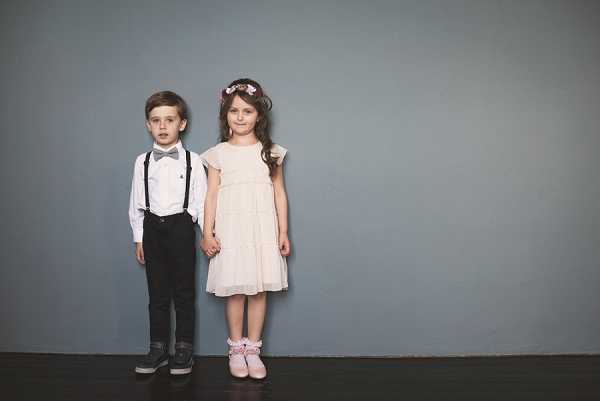 Two young children, likely a ring bearer and flower girl, pose holding hands against a plain muted blue-grey wall. The boy wears black trousers, a white dress shirt, grey suspenders, and a grey bow tie with dark sneakers, while the girl wears a tiered cream/blush tulle dress with cap sleeves, pink ballet flats, and a floral crown with small pink blooms. The portrait is a full-length shot with a clean, minimal backdrop giving it a editorial feel. The styling is classic with soft, muted tones throughout.