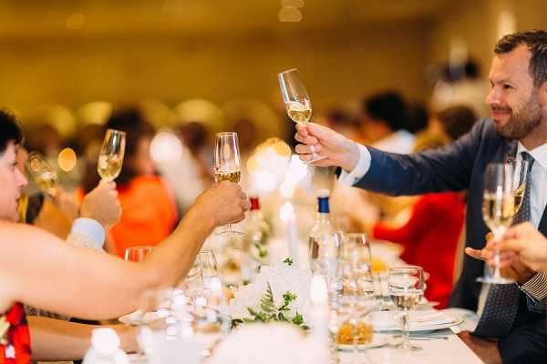 Guests are raising champagne flutes in a toast during a wedding reception dinner, set in a warmly lit indoor ballroom or event hall. The foreground shows a decorated dining table with a low centerpiece of white flowers and greenery, small votive candles, wine glasses, and a wine bottle. A man in a navy suit with a tie is prominently visible on the right side, smiling as he joins the toast, while other guests in colorful attire — including orange and red outfits — are visible in the background, which is softly blurred with warm golden bokeh lighting. The shot is taken at table level, creating a close-up, immersive perspective of the toasting moment.