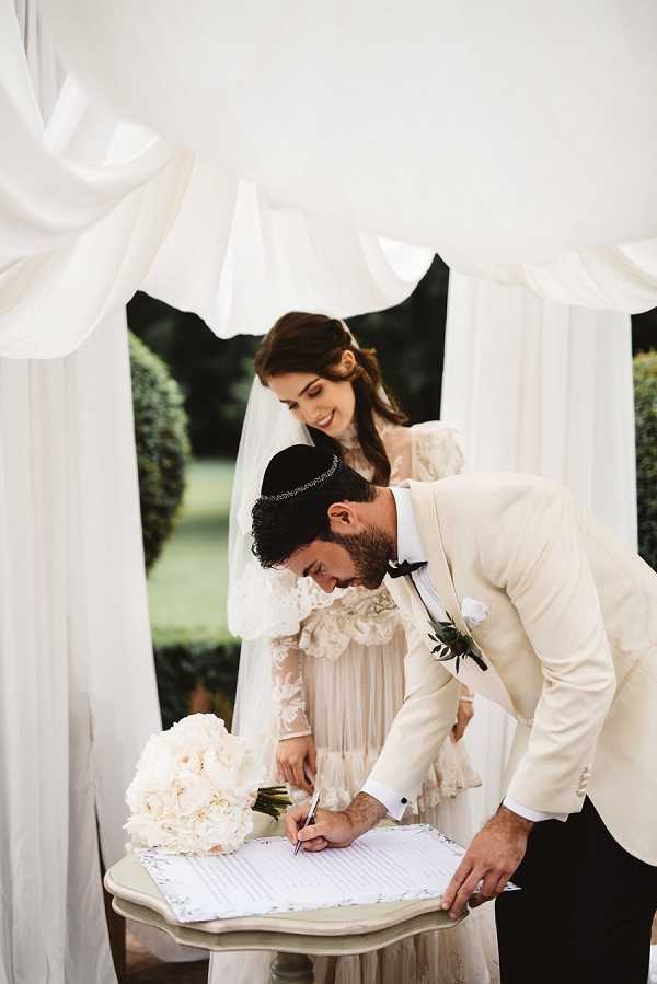 The groom signs what appears to be a ketubah (Jewish marriage contract) on a small ornate table during an outdoor ceremony, while the bride stands beside him smiling. The groom wears a cream blazer with black trousers, a black bow tie, a white boutonniere with dark foliage, and a kippah with subtle embellishment. The bride wears a romantic ivory lace and tulle gown with ruffled details and long sleeves, paired with a veil and a delicate headpiece. A large bouquet of ivory and blush peonies rests on the table beside the document. The couple stands beneath a chuppah draped with flowing white fabric panels, with manicured topiary hedges visible in the background, indicating a formal garden setting. The shot is a medium portrait framing both figures from the waist up.