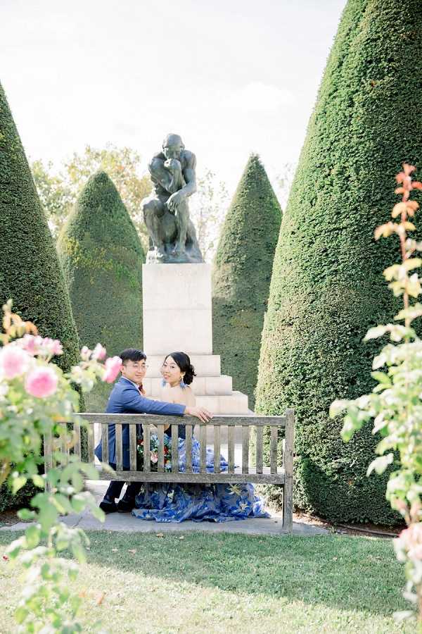 A couple poses seated on a wooden garden bench in an outdoor formal garden setting, with Rodin's 'The Thinker' bronze sculpture on its stone pedestal visible directly behind them. The groom wears a navy blue suit with a burgundy tie, while the bride wears a striking blue floral-printed ball gown with an off-shoulder neckline featuring blue tassel or feather earring details. Cone-shaped trimmed topiary hedges frame the couple symmetrically on both sides, and pink roses are visible in the foreground, slightly out of focus. This is a medium-distance portrait shot with the couple relaxed and leaning into each other, captured in a classic garden style with soft natural light.