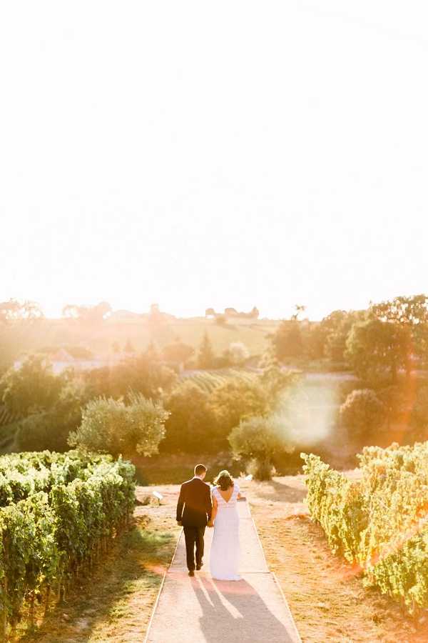 A couple portrait taken outdoors in a vineyard setting at golden hour, with the bride and groom walking away from the camera along a narrow path flanked by rows of grapevines. The groom wears a dark suit and the bride wears a white gown with an open V-back and lace detailing. Strong warm backlighting creates pronounced lens flare and a glowing haze across the scene. Rolling hills and trees are visible in the background. Wide shot composition with the couple centered in the lower third of the frame.