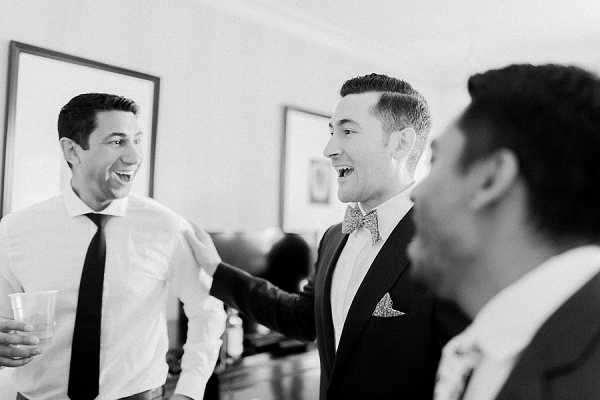 A black-and-white getting-ready shot capturing three men laughing together in an indoor room, likely a hotel or private residence. The central figure, presumably the groom, is dressed in a dark suit jacket with a patterned bow tie and a pocket square, while the man to his left wears a white dress shirt with a dark tie and holds a glass. The third man is partially visible in the foreground, out of focus. The image has strong contrast with bright highlights and deep shadows, conveying a candid, joyful moment among the groom and groomsmen. Close-to-medium portrait composition with a shallow depth of field.