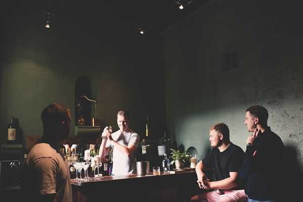 A cocktail hour scene set at an indoor bar with dark green walls and moody, low-level track lighting overhead. A bartender in a white t-shirt is shaking a cocktail behind the wooden bar counter, which is lined with glassware, bottles, and small potted plants. Three male guests — one with his back to the camera in a tan shirt, and two seated at the bar in dark shirts with one wearing light pink trousers — are gathered at the bar chatting. The overall decor style is modern and intimate, with a dark, muted color palette. Medium wide shot capturing the full bar scene and interaction between the bartender and guests.
