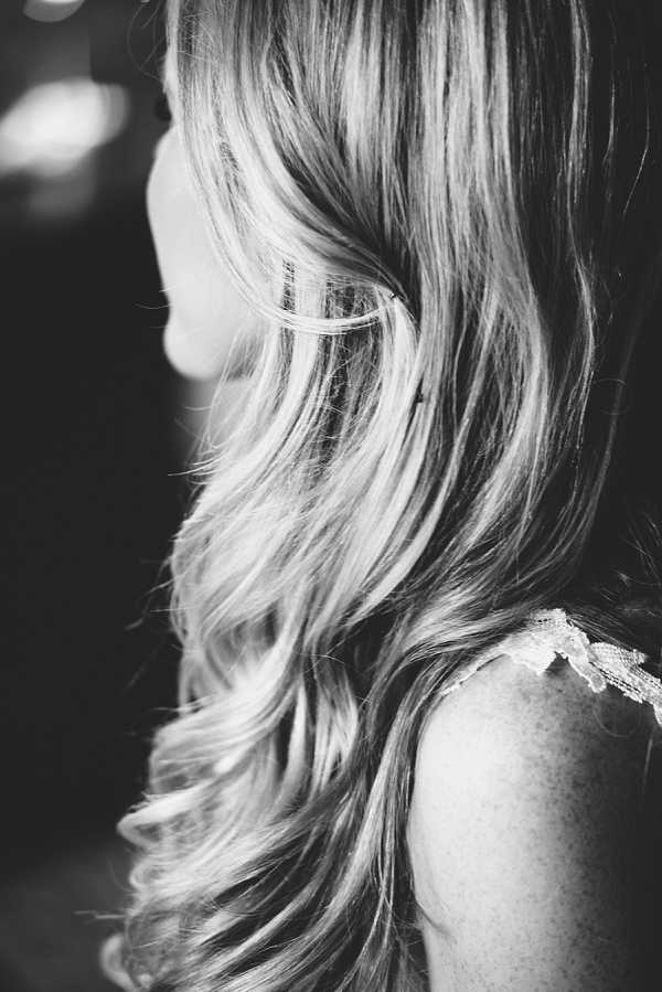 Black-and-white close-up portrait of a bride during the getting-ready phase, shot from behind and slightly to the side. The image focuses on her long, loosely curled hair, which falls over her shoulder in soft waves with high contrast between light and dark tones. A lace strap of what appears to be a bridal gown or lingerie is visible at her shoulder. The background is dark and blurred, keeping all attention on the hair styling detail.