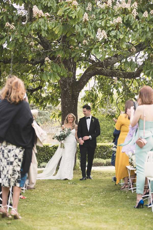 A newly married couple walks back up the aisle following an outdoor ceremony held on a garden lawn, with guests standing and applauding on either side. The bride wears a fitted, lace white gown with a long train and carries a loose bouquet of greenery and white blooms, while the groom is dressed in a black tuxedo with a black bow tie and a small white buttonhole. Guests are seated and standing on white folding chairs along both sides of the grass aisle, dressed in a mix of mustard yellow, mint green, and lavender outfits. The shot is a medium wide portrait taken from behind the guests, framing the couple between the two rows of attendees with a large flowering tree visible in the background.