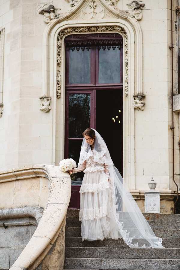 A bridal portrait taken outdoors on the stone steps of a chateau or grand historic building, with ornate carved limestone facade and a large dark burgundy-framed arched doorway visible behind the bride. The bride stands alone on the steps, looking down with a slight smile, wearing a distinctive tiered tulle and lace wedding gown in ivory with blush undertones, featuring ruffled layers and long lace-trimmed sleeves. She wears a cathedral-length veil with lace edging and holds a compact round bouquet of white peonies or garden roses. The composition is a full-length portrait shot, capturing the architectural detail of the building alongside the bride's styling. Potential venue feature image.