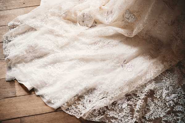 Close-up detail shot of an ivory lace wedding dress laid out on a rustic wooden plank floor. The gown features intricate floral and botanical lace patterns across multiple layers, with a scalloped lace hem visible at the bottom edge. Natural light falls across the fabric, highlighting the texture and delicate embroidery details of the overlay. No people are visible; this is a getting-ready detail image focused entirely on the dress.