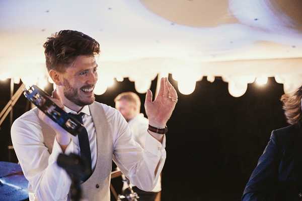 A man, likely a wedding guest or groomsman, is dancing and playing a tambourine during what appears to be an indoor evening reception. He is wearing a light beige suit jacket, white dress shirt, and dark navy tie, with his jacket open and sleeves partially rolled, suggesting the party is in full swing. The setting appears to be a marquee or tent reception space, with warm Edison-style bulb lighting visible along the tent edges in the background. The shot is a mid-length portrait with another guest in a navy suit partially visible at the right edge, and a second person visible in the background.