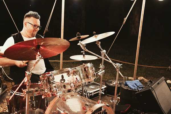 A male drummer performs live at an outdoor evening wedding reception, playing a clear acrylic drum kit with red/bronze cymbals under a white tent structure. The musician is wearing glasses, a white shirt, and a dark vest, and is actively mid-play with drumsticks raised. The setting is dimly lit with artificial lighting illuminating the performer and kit against a dark background, suggesting a nighttime outdoor reception entertainment setup. The shot is a medium close-up focused on the performer and drum kit from a slight low angle.