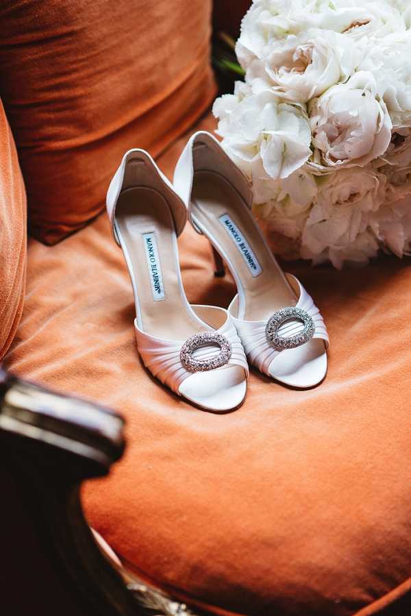 A close-up detail shot of a pair of white satin Manolo Blahnik peep-toe heels featuring crystal embellished circular buckle ornaments at the toe, placed on a burnt orange velvet chair seat. Partially visible in the upper right corner is a bridal bouquet composed of white and blush peonies and hydrangeas. The styling is classic and polished, with the warm terracotta tone of the velvet upholstery contrasting against the white satin shoes.