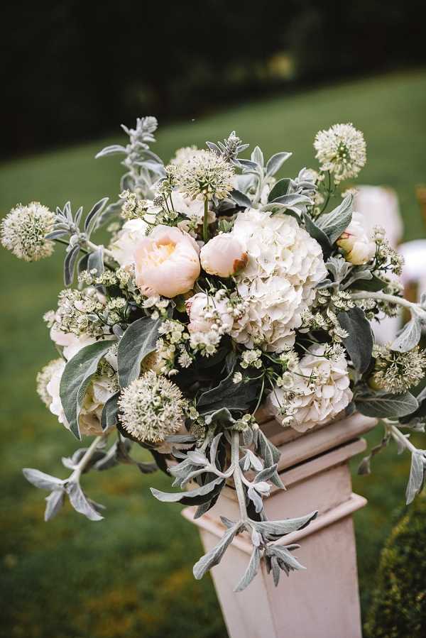 Close-up detail shot of a ceremony floral arrangement displayed on a pale blush pedestal column at an outdoor wedding. The arrangement features white hydrangeas, blush peonies, small white allium-style globe flowers, delicate white filler blooms, and generous amounts of silver-green lamb's ear foliage trailing over the sides of the pedestal. The color palette is soft blush and white with silvery-grey green foliage, consistent with a classic romantic styling. The background is a blurred green lawn, indicating a garden ceremony setting.