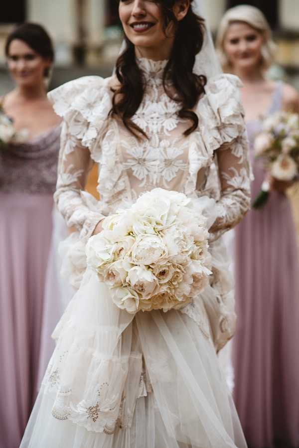 A portrait-style close-up of a bride holding a large rounded bouquet of ivory and blush peonies and garden roses, with soft tulle ribbon trailing below. The bride wears a distinctive long-sleeve, high-neck wedding gown with sheer lace appliqué detailing, ruffled shoulders, and a tiered tulle skirt with lace trim — a romantic, vintage-inspired silhouette. Her dark hair is worn down in loose waves and she has a veil. Two bridesmaids in floor-length dusty mauve/lilac dresses are visible but out of focus in the background, one also holding a small bouquet. The setting appears to be outdoors in front of a building. The styling is classic with vintage influences.