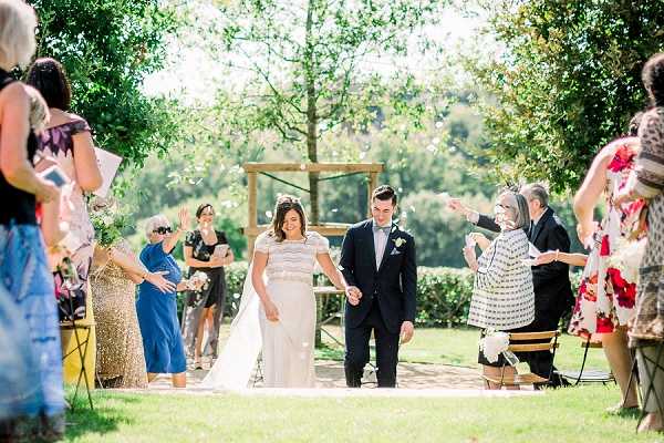 A newly married couple walks back down the aisle following an outdoor ceremony, with guests on both sides tossing confetti. The bride wears a long ivory dress with a beaded or embroidered cap-sleeve bodice, and the groom wears a navy suit with a light blue pocket square and a white boutonniere. The ceremony took place in a garden setting with a simple rustic wooden arch visible in the background. Approximately 10–12 guests are visible lining the aisle, dressed in a mix of colorful outfits including a gold sequined dress and a floral-patterned dress. The overall styling is classic and relaxed, and the image is a wide shot taken in bright natural daylight.