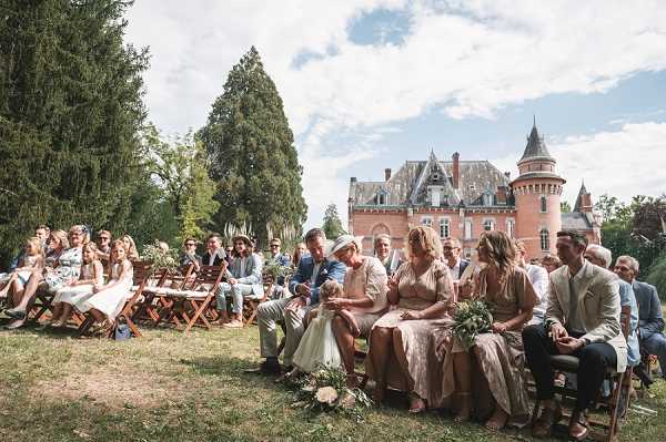An outdoor wedding ceremony taking place on the grounds of a French chateau, with the pink-brick castle building and its distinctive turret visible in the background. Approximately 30-40 guests are seated on wooden folding chairs arranged in rows on a lawn, all facing toward the ceremony. In the foreground, front-row guests include women wearing gold and champagne-toned dresses, likely bridesmaids or family members, and a small flower girl in a white dress standing near the seated guests. The overall styling is classic with warm, neutral tones — gold, beige, and cream — visible in the guest attire, and a loose greenery and white floral arrangement is visible at the end of a row. The shot is a wide angle taken from the side of the seating area, capturing both the audience and the chateau backdrop. Potential venue feature image.