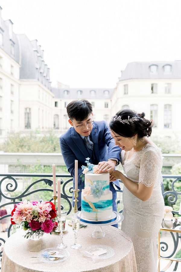 A couple cuts their wedding cake on an outdoor balcony with Haussmann-style Parisian buildings visible in the background. The groom wears a navy blue suit with a patterned tie and glasses, while the bride wears a short-sleeve lace fitted gown and a delicate crystal hair vine with her hair in an updo. The two-tier cake features a watercolor blue and teal design with peach and blue sugar flowers on a white base with a navy blue base board. The round table is dressed in a champagne sequined linen and holds two blush taper candles in gold candlesticks, crystal champagne flutes, and a floral arrangement of hot pink, red, and blush dahlias and garden roses. The shot is a medium portrait framing both subjects from the waist up.