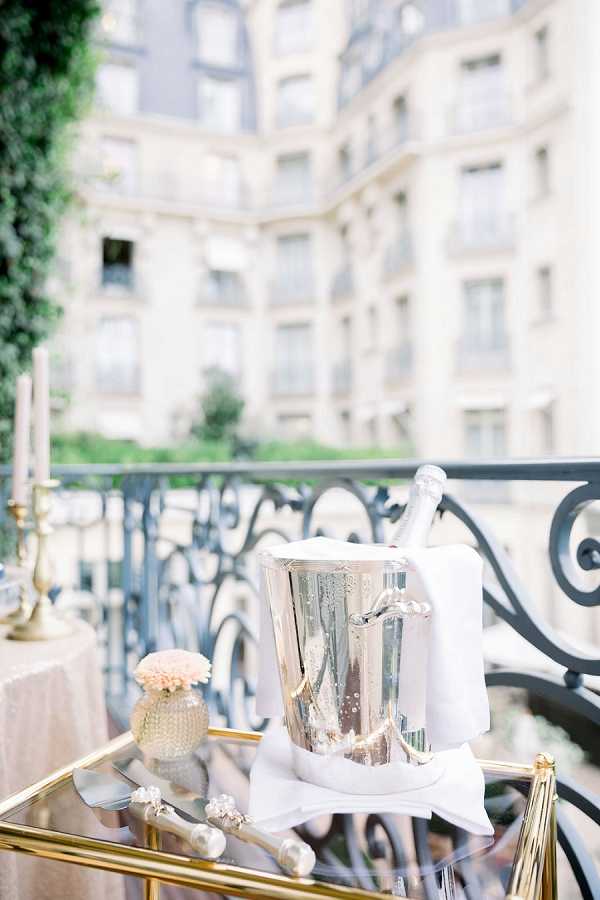 A detail close-up shot of a gold-framed glass bar cart positioned on an outdoor balcony with ornate wrought iron railings, set against a blurred Haussmann-style Parisian building facade in the background. On the cart sits a polished silver champagne ice bucket holding a bottle, draped with a white linen cloth, alongside a small vase with a single blush carnation or ranunculus bloom and a decorative cake server with pearl-style handle details. To the left, tall taper candles in gold brass candlestick holders are partially visible beside what appears to be a blush or champagne-toned linen table. The styling palette is soft blush, ivory, and gold with classic Parisian details, suggesting a refined cocktail hour or reception setup. Potential venue feature image.