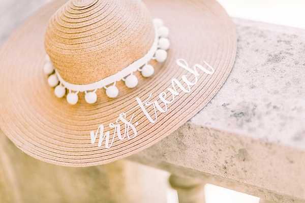 Close-up detail shot of a wide-brim straw floppy hat personalized with the text 'mrs french' in white script lettering on the brim, trimmed with a white pom-pom ribbon band. The hat is resting on a stone surface, likely a balustrade or ledge. The accessory has a relaxed, playful bridal style and appears to be a getting-ready or bridal detail prop.