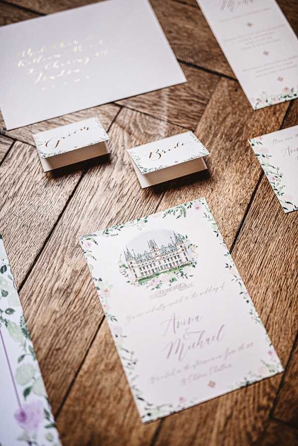 A flat lay detail shot of a wedding stationery suite arranged on a herringbone oak wood surface. The collection includes a wedding invitation featuring a watercolor illustration of a French chateau surrounded by a floral border in soft blush pink, sage green, and ivory tones, with calligraphy script reading 'Anna & Michael.' Also visible are two small favor or place card boxes labeled 'Groom' and 'Bride' in gold calligraphy, a white envelope with gold foil hand-lettered addressing, a menu or order of service card, and additional stationery pieces partially visible at the edges. The overall design palette is blush, white, sage, and gold, with a romantic watercolor floral motif consistent throughout the suite.