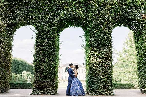 A couple poses for a portrait in the formal gardens of a French chateau, standing between tall ivy-covered hedge arches that frame the shot. The bride wears a floor-length blue and white floral ballgown, and the groom is dressed in a navy suit; they face each other in an embrace with a white bouquet visible. A classical chateau building is visible in the background through the central arch, indicating a historic estate venue. The image is a wide shot with the architectural hedge columns creating strong vertical symmetry around the couple. Potential venue feature image.