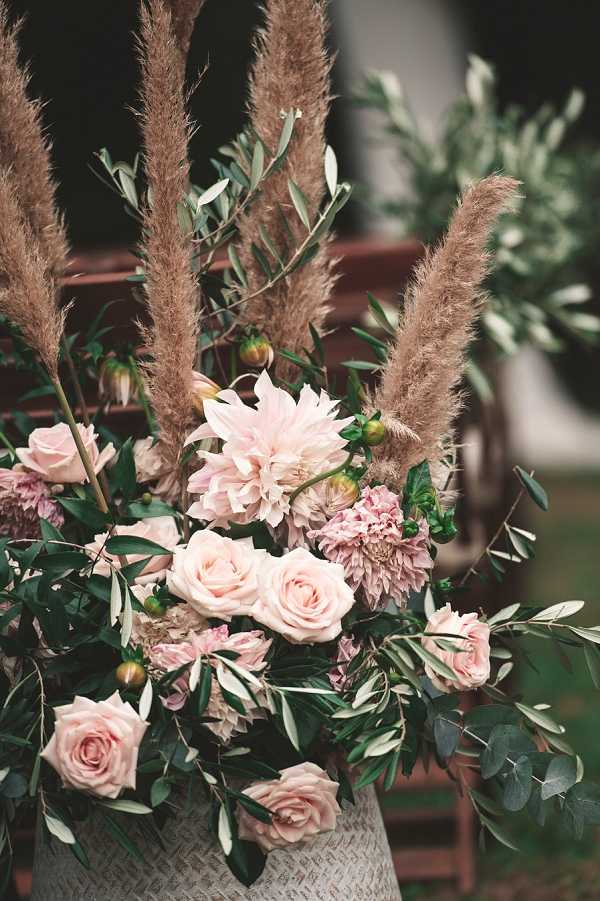 Close-up detail shot of a ceremony aisle floral arrangement placed in a white woven basket, positioned beside a wooden chair with additional arrangements visible in the blurred background. The arrangement features blush pink roses, blush and dusty pink dahlias with green buds, and tall pampas grass plumes extending upward, styled in a boho aesthetic. Olive branches and eucalyptus provide lush greenery throughout the composition. The overall palette is blush pink, dusty rose, and green with warm tan tones from the pampas grass.