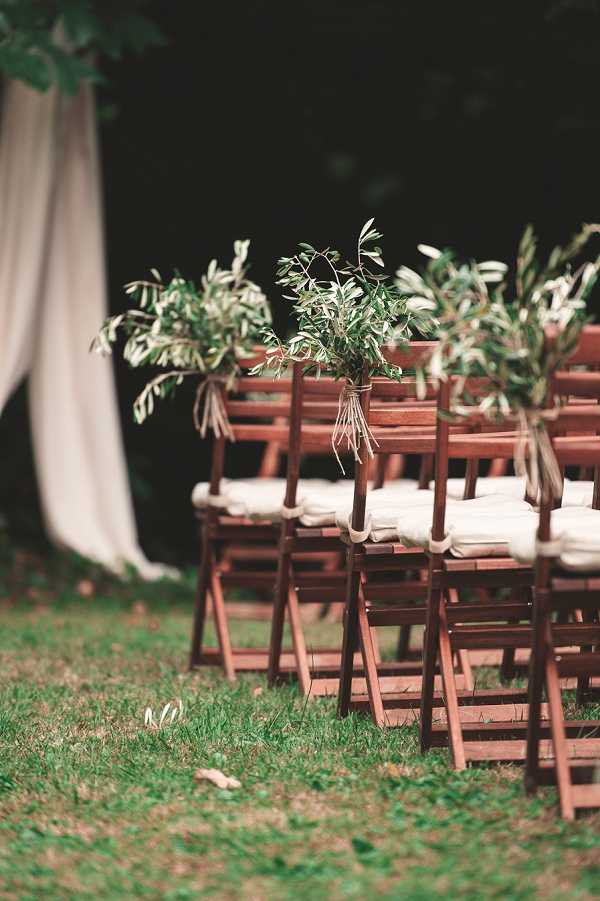 Detail shot of an outdoor wedding ceremony setup on a grass lawn, showing rows of dark wood folding chairs with ivory cushions. The aisle chairs are decorated with small bunches of olive branches tied with twine or thin ribbon, giving a rustic, natural aesthetic. A draped ivory or white fabric panel is partially visible on the left side of the frame, likely part of a ceremony arch or backdrop. No guests are present, suggesting this was taken before the ceremony. The composition is a shallow-depth-of-field portrait-orientation shot with the foreground chair in focus and the remaining rows softly blurred.