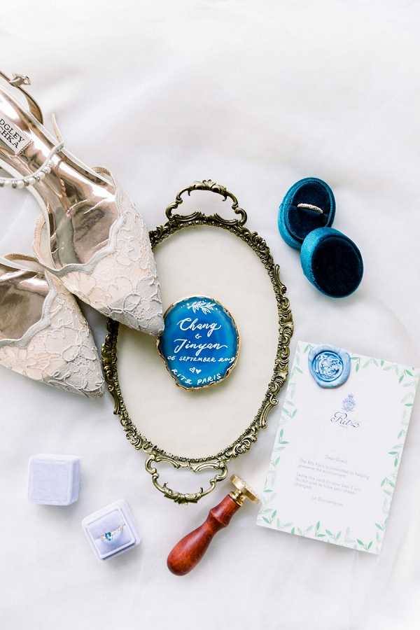 A flat lay detail shot of wedding accessories arranged on a white surface, centered around an ornate gold oval vanity tray. Inside the tray sits a teal and gold hand-lettered wedding disc reading 'Chang & Jinyan, 06 September 2019, Ritz Paris.' Surrounding the tray are: a pair of silver strappy heeled sandals with lace detailing branded Badgley Mischka, a teal velvet double ring box holding a wedding band, a small lavender velvet ring box with a gemstone ring inside, a red wood-handled wax seal stamp, and a white Ritz Paris stationery card with green botanical illustrated borders sealed with a pale blue wax seal. The overall color palette features teal, gold, lavender, and ivory with a classic Parisian aesthetic.