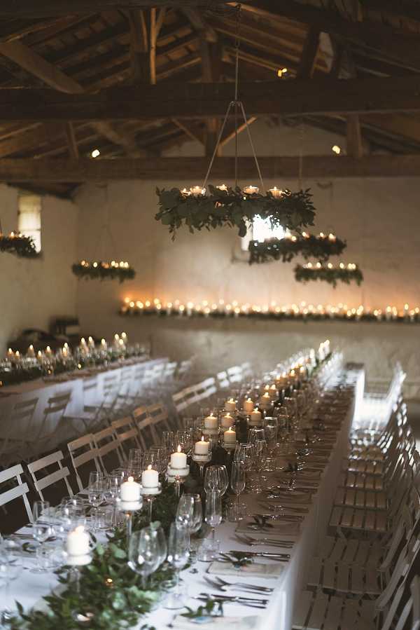 A reception dinner setup inside a rustic barn or stone outbuilding with exposed wooden beam ceiling and raw stone walls. Multiple long banquet tables are dressed with white linen and styled with a greenery runner featuring trailing eucalyptus or similar foliage, white pillar candles on varying-height candlestick holders, and full glassware and silverware place settings. Overhead, rectangular hoops wrapped in greenery and fitted with tealight candles hang from the ceiling as chandeliers, and a continuous row of tealights lines a ledge along the back wall, creating warm candlelit ambient lighting throughout the space. The overall styling is rustic-organic, with an all-greenery and white candle palette and no floral color. Wide interior shot taken from one end of the room looking down the length of the tables.