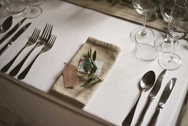 Close-up detail shot of a wedding reception place setting on a white linen tablecloth. The setting includes silver cutlery arranged with two forks on the left and a spoon and knife on the right, alongside multiple clear wine and water glasses. A natural burlap/linen napkin is folded at the center and topped with a small sprig of eucalyptus, a miniature gift or favor box, and a terracotta-colored luggage-style place card tag with handwritten calligraphy. The overall styling is minimal and rustic-natural, with a neutral palette of white, natural linen, and silver.