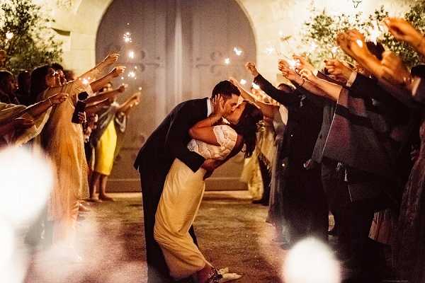 A couple shares a dip kiss during a sparkler send-off at an outdoor evening reception, with approximately 20 guests lining both sides holding lit sparklers raised overhead to form an arch. The bride wears a two-piece or separates-style ivory and yellow wedding dress with a lace or embellished bodice, while the groom is dressed in a dark navy or black suit. The warm glow of the sparklers illuminates the scene against a nighttime backdrop featuring a stone arched doorway and manicured hedges or topiaries. The shot is taken at ground level from a wide portrait perspective, with soft bokeh from nearby sparklers framing the foreground.