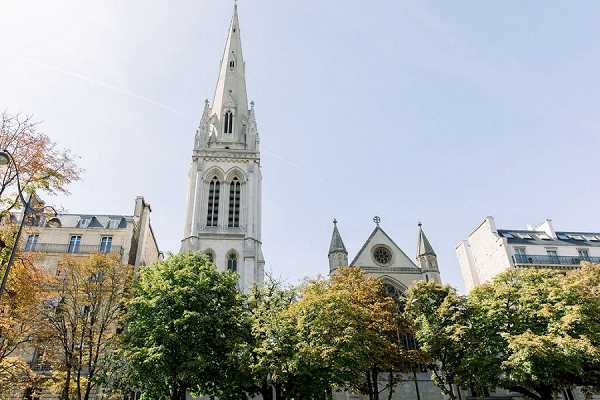 Wide-angle exterior shot looking upward at a white Gothic-style church with a tall pointed spire, rose window, and ornate stone detailing, set within a Parisian urban streetscape. The church is partially framed by mature trees and flanked by classic Haussmann-style apartment buildings on either side. No people are visible in the frame. The image appears to be a venue establishing shot taken in natural daylight. Potential venue feature image.