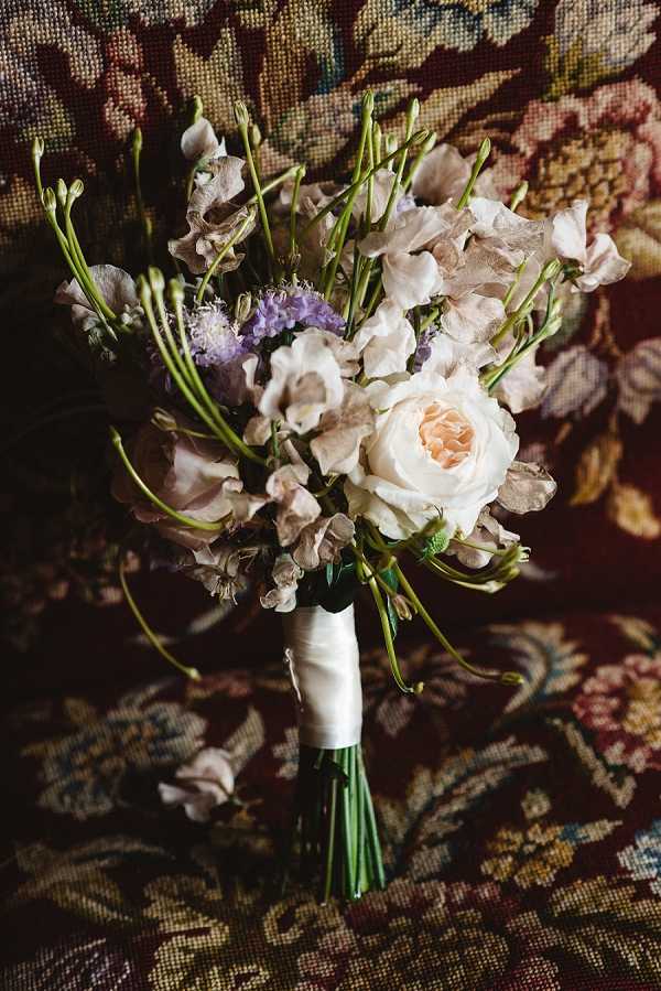Close-up detail shot of a bridal bouquet resting against a richly patterned tapestry or upholstered fabric in deep burgundy, gold, and green floral motifs. The bouquet features a cream garden rose as the focal bloom, surrounded by blush and taupe sweet peas, lavender statice, and trailing green stems with budding flowers, giving it a loose, wild, garden-gathered aesthetic. The stems are bound with a white satin ribbon. The overall color palette is soft and muted — cream, blush, taupe, and lavender — contrasting against the dark, ornate background fabric.