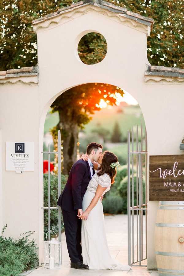 A bride and groom share a kiss while standing in an arched stone gateway at what appears to be a French vineyard or domaine, photographed during golden hour with warm sunset light glowing through the arch behind them. The bride wears a flowing ivory lace gown with short sleeves and has white floral hair accessories, while the groom wears a navy suit; they are holding hands during the kiss. A wooden welcome sign with white script reading 'Welcome Maja & [name] August 18' is visible to the right, along with a wine barrel, a white lantern on the ground, and a 'Visites' information placard on the left pillar. This is a full-length portrait shot with the couple centered in the archway, styled with a romantic rustic-chic aesthetic consistent with a French wine estate setting. Potential venue feature image.