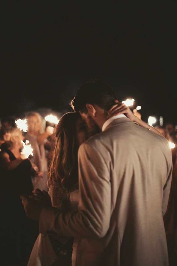 A couple shares a kiss during a nighttime sparkler send-off, surrounded by a crowd of approximately 20 or more guests holding lit sparklers. The groom wears a light tan or beige suit, and the bride wears a long-sleeved white lace dress with her hair down. The sparklers create warm, glowing light points in the dark background, producing a dramatic contrast between the illuminated couple in the foreground and the near-black night sky. The image is a close portrait shot taken from behind and slightly to the side of the couple, with guests softly out of focus in the background.