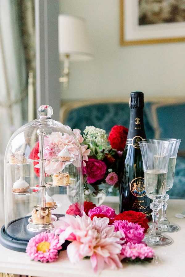 A close-up detail shot of a getting-ready or welcome amenity display set on a marble side table in what appears to be a hotel suite, with a teal upholstered sofa visible in the background. A glass cloche covers a tiered silver stand holding small pastries and petit fours, accompanied by a bottle of Ritz Paris Reserve Champagne Brut and two crystal champagne flutes, one partially filled. Loose cut flowers in hot pink, magenta, and red tones — including dahlias, peonies, and ranunculus — are scattered around the base of the display alongside a fuller floral arrangement behind the bottle. The overall styling palette is bold and romantic with deep jewel-toned florals against the soft blue-grey tones of the room.
